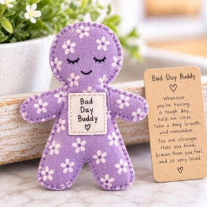 May include: A purple fabric doll with a white floral pattern, featuring embroidered closed eyes and a smile. A square patch on the chest reads Bad Day Buddy with a heart. Beside it is a rectangular card with the text Bad Day Buddy and a poem.