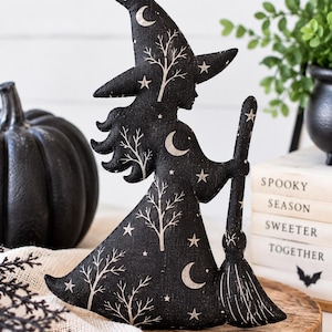May include: A black fabric witch figurine featuring a white printed pattern of crescent moons, stars, and bare trees. The witch wears a pointed hat and holds a broomstick. The fabric has a textured appearance, and the design is flat and cutout.