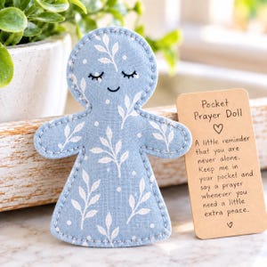 May include: A light blue fabric doll with a white leaf and dot pattern, featuring embroidered closed eyes and a small smile. Beside it is a rectangular tan card with the text: Pocket Prayer Doll, A little reminder that you are never alone. Keep me in your pocket and say a prayer whenever you need a little extra peace.