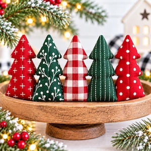 May include: Five plush fabric Christmas tree ornaments arranged in a row on a wooden pedestal. The trees feature various patterns including red with white plus signs, green with white sprigs, red and white gingham, green with white dashes, and red with white polka dots.