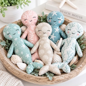 May include: Five plush mermaid dolls with embroidered faces, each featuring a unique fabric pattern including starfish, seashells, and coral motifs in shades of pink, blue, teal, and beige, arranged together in a round wooden bowl.