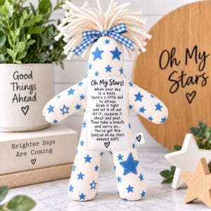 Pode incluir: Uma boneca antiestresse de tecido cor de creme com um padrão de estrelas azuis, finalizada com cabelo de fio creme e um laço xadrez azul. Um remendo retangular branco no peito exibe o texto: Oh My Stars! When your day is a mess...
