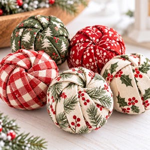 Op de afbeelding: Een set van vijf met stof beklede bolvormige ornamenten met diverse feestelijke patronen, waaronder rood-wit gingham, groen met witte dennenbomen, rood met witte bessen en crème met hulstbladeren en rode bessen.