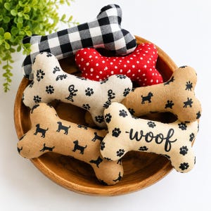 Può includere: Una collezione di cinque giocattoli in tessuto a forma di osso per cani in una ciotola di legno. I giocattoli presentano vari motivi, tra cui scacchi bianchi e neri, rosso a pois bianchi e beige con impronte di zampe e la parola woof.