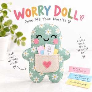 Puede incluir: Una muñeca de tela verde con estampado de margaritas blancas, que presenta un bolsillo beige con un corazón rosa, mejillas rosadas y ojos bordados. La muñeca sostiene pequeños papeles con textos como "You Got This" y "Be Kind to Yourself".