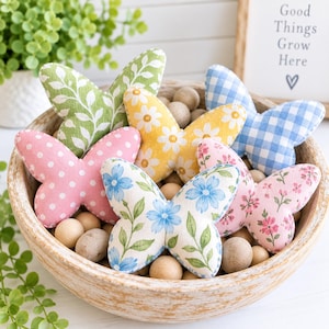 May include: A collection of six fabric butterfly ornaments in a wooden bowl filled with wooden beads. The butterflies feature various patterns including green leaves, yellow daisies, blue gingham, pink polka dots, and floral prints.
