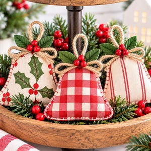 May include: Three fabric bell ornaments featuring festive patterns: one with green holly leaves and red berries, one with a red and white gingham check, and one with red vertical stripes. Each bell has a jute twine loop and a decorative red berry accent.