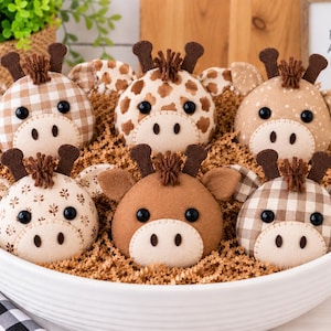 May include: A collection of six round plush giraffe heads in a white bowl filled with brown paper shreds. Each plush features black button eyes, a cream-coloured muzzle, brown felt horns, and a small tuft of brown yarn hair in various fabric patterns.