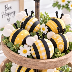 May include: A collection of spherical bee ornaments featuring yellow and black patterns, including gingham, polka dots, and floral prints. Each bee has white felt wings and black wire antennae, arranged on a tiered wooden tray with greenery.