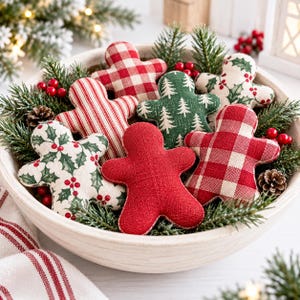 May include: A collection of six fabric gingerbread-shaped ornaments nestled in a round wooden bowl. The ornaments feature various patterns including red and white gingham, red and white stripes, solid red, green with white trees, and cream with holly.