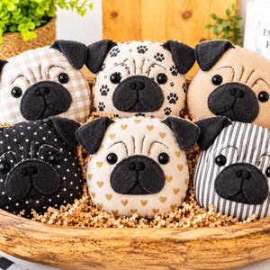 Könnte beinhalten: Sechs Mops-Köpfe aus Plüsch in einer Holzschale. Die Möpse haben Ohren und Nasen aus schwarzem Filz, Kunststoffaugen und verschiedene Stoffmuster, darunter beiges Vichy-Karo, Pfotenabdrücke, einfarbiges Beige, schwarze Punkte, goldene Herzen und graue Streifen.