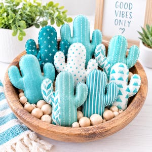 Op de afbeelding: Een verzameling pluche cactusornamenten in diverse tinten blauwgroen, turquoise en wit, met patronen zoals strepen, stippen, kruisjes en bladeren. Deze stoffen cactussen zijn gerangschikt in een ronde houten kom met houten kralen.