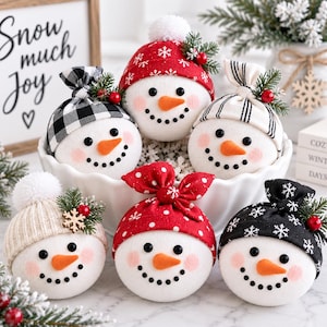 Op de afbeelding: Zes ronde sneeuwman-ornamenten met witte stoffen gezichten, oranje wortelneuzen en zwarte kraaloogjes en mondjes. Elke sneeuwman draagt een unieke stoffen muts, inclusief rode, zwarte en witte patronen, versierd met dennentakjes en rode bessen.