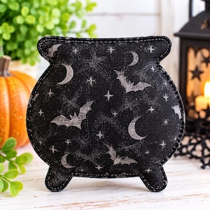 May include: A black fabric cauldron-shaped ornament featuring a repeating pattern of grey crescent moons, stars, and flying bats. The item has a stitched border and three small feet at the base, crafted from a textured material with a dark finish.