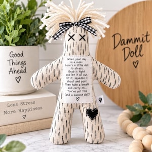 Puede incluir: Una muñeca de tela beige con patrones de rayas negras, cabello de hilo blanco y un lazo de cuadros vichy en blanco y negro. Un parche blanco en el pecho dice: When your day is a mess, here's a little doll to stress. Grab it tight.