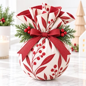 May include: A round fabric pouch featuring a cream-colored base with a red botanical print of leaves and berries. The top is gathered and tied with a solid red ribbon, accented by two sprigs of green pine needles and clusters of red berries.
