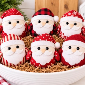 May include: Six plush Santa Claus ornaments with round bodies, white felt beards, and hats in various patterns including red polka dots, black and red buffalo plaid, red snowflakes, red stripes, and solid red, all nestled in a white bowl.