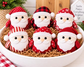 May include: Six plush Santa Claus ornaments with round bodies, white felt beards, and hats in various patterns including red polka dots, black and red buffalo plaid, red snowflakes, red stripes, and solid red, all nestled in a white bowl.
