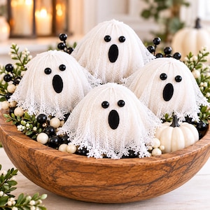 May include: Four ghost figures crafted from white lace fabric with black oval eyes and mouths, arranged in a large wooden bowl filled with black and white decorative berries and a small white pumpkin, creating a festive display.