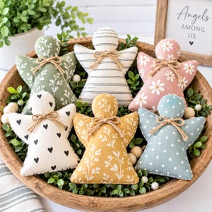 May include: A set of six fabric angel ornaments in a wooden bowl. Each angel features a unique pattern including white floral on sage green, black stripes on cream, white daisies on pink, black hearts on cream, white floral on mustard, and white polka dots on blue. Each angel is accented with a small jute twine bow at the neck.