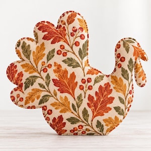 May include: A turkey-shaped decorative cushion featuring a cream-coloured fabric base with a printed pattern of autumn leaves in shades of orange, red, and brown, interspersed with small red berries and green foliage, finished with a piped edge.