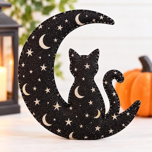Op de afbeelding: Een zwart stoffen ornament in de vorm van een maansikkel met een geprint patroon van kleine beige sterren en maansikkels. Een zwart kattensilhouet is in het midden van de maan verwerkt, met de staart omhoog krullend langs de onderrand.
