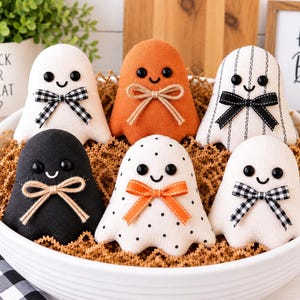 Può includere: Sei figure di fantasmi in peluche in una ciotola bianca, ognuna con occhi a bottone neri, un sorriso ricamato e un fiocco decorativo. I fantasmi includono tessuto bianco, arancione e nero, oltre a tessuto bianco con righe o pois neri.