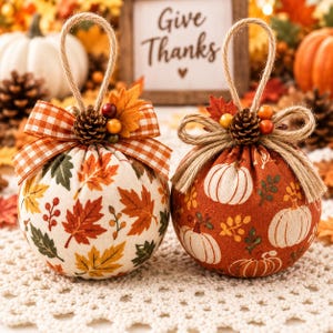 May include: Two round fabric ornaments with jute hanging loops. The left ornament features a cream background with orange, yellow, and green autumn leaves and an orange gingham bow. The right ornament has a rust background with white pumpkins.