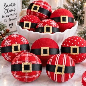 Könnte beinhalten: Eine Sammlung runder roter Weihnachtskugeln mit verschiedenen Mustern wie Karos, Punkten, Schneeflocken und Streifen. Jede Kugel ist mit einem schwarzen Band und einer quadratischen, gold glitzernden Schnalle als Gürtel verziert.