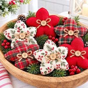 May include: A collection of five fabric flower ornaments in a wooden bowl, featuring red felt, red and green plaid, and white fabric with red berry patterns. Each flower is centered with a light brown wooden button and twine bow.