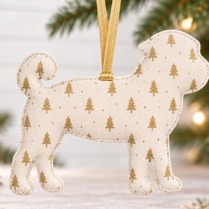 Pode incluir: Um enfeite de Natal de tecido em forma de cão, na cor branco-sujo, com um padrão repetitivo de pequenos pinheiros dourados metalizados. O enfeite está suspenso por uma fita dourada fina presa na parte superior central das costas.
