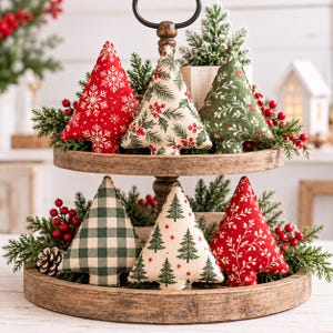 May include: A two-tiered wooden tray holding six fabric Christmas tree ornaments. The trees feature red snowflake, cream holly, green foliage, green buffalo check, cream pine tree, and red vine patterns, accented with faux greenery and red berries.