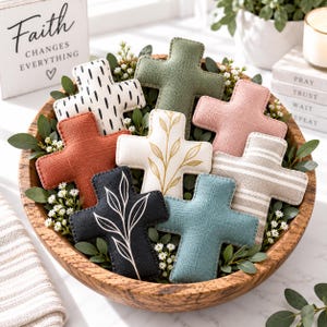 May include: A collection of nine fabric crosses in a wooden bowl, featuring various colours and patterns including solid olive green, terracotta, dusty pink, teal, black with white leaf prints, white with black dashes, and white with gold leaf designs.