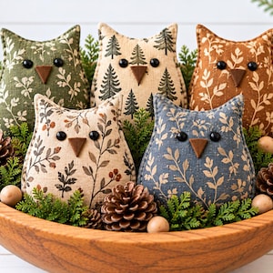 May include: Five stuffed fabric owl ornaments in a wooden bowl. The owls feature various patterns including green, beige, and blue with leaf, berry, and pine tree prints. Each owl has two black bead eyes and a brown felt triangular beak.