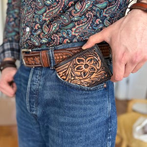 May include: A brown leather wallet with intricate floral tooling, tucked into the front pocket of blue jeans.