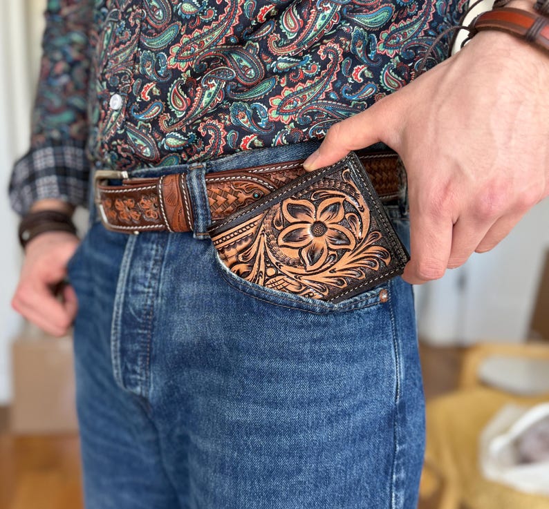 May include: A brown leather wallet with intricate floral tooling, tucked into the front pocket of blue jeans.