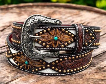 May include: A brown leather belt with a white leather inlay and a silver buckle. The belt is decorated with gold studs and floral designs. The buckle has a turquoise stone in the center.