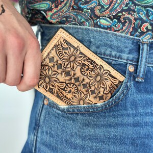 May include: A brown leather wallet with a floral design is tucked into the front pocket of a pair of blue jeans.
