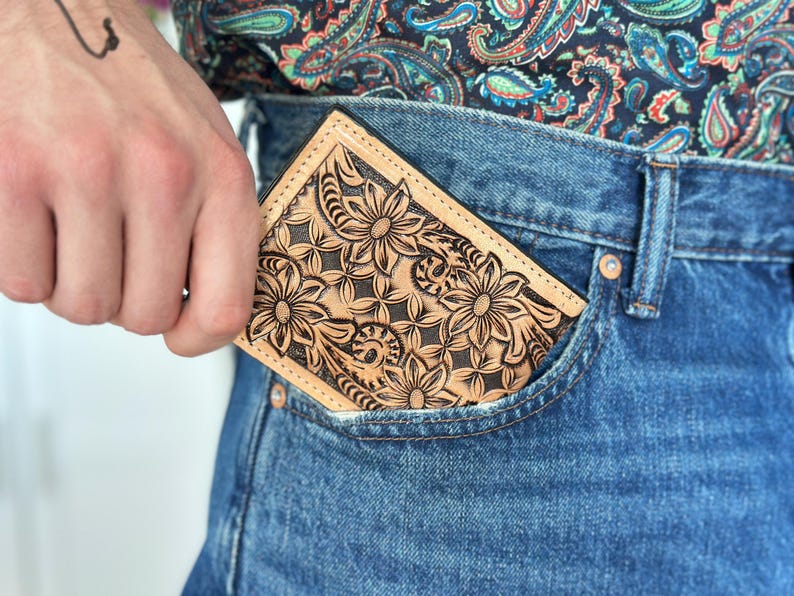 May include: A brown leather wallet with a floral design is tucked into the front pocket of a pair of blue jeans.