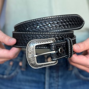 May include: A black leather belt with a woven pattern and a silver buckle with an ornate design. The belt has white stitching around the edges.