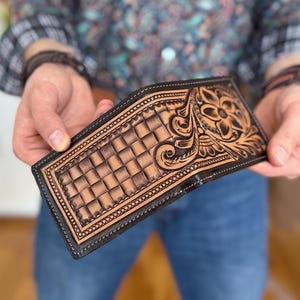 May include: A brown leather wallet with a detailed floral design and a checkered pattern. The wallet has a black border and stitching.