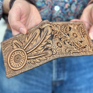 May include: A brown leather wallet with intricate tooled designs of feathers and flowers. The wallet is closed and has a dark brown edge.
