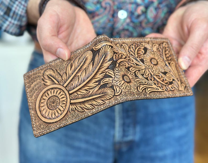 May include: A brown leather wallet with intricate tooled designs of feathers and flowers. The wallet is closed and has a dark brown edge.