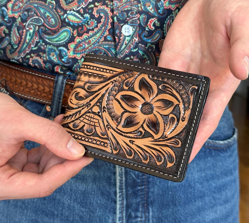 May include: A brown leather wallet with a tooled floral design. The wallet has a black stitching around the edges.