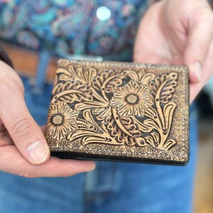 May include: A brown leather wallet with a floral tooled design. The wallet has a black border and is closed.