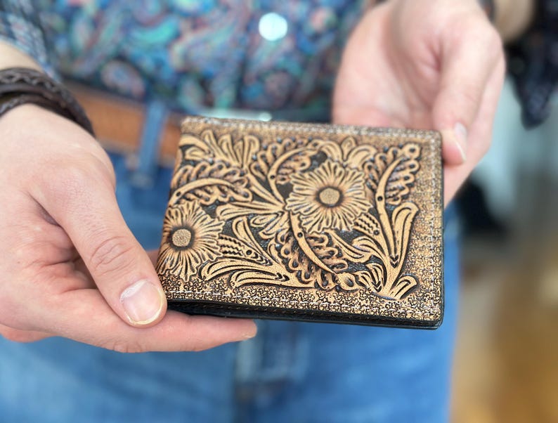 May include: A brown leather wallet with a floral tooled design. The wallet has a black border and is closed.