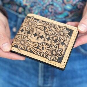 May include: A brown leather wallet with a floral design embossed on the front. The wallet has a black stitching around the edges.