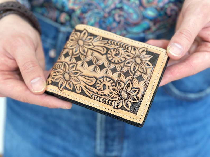 May include: A brown leather wallet with a floral design embossed on the front. The wallet has a black stitching around the edges.