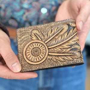 May include: A brown leather wallet with a tooled design of feathers and a dreamcatcher. The wallet has a black stitching around the edges.