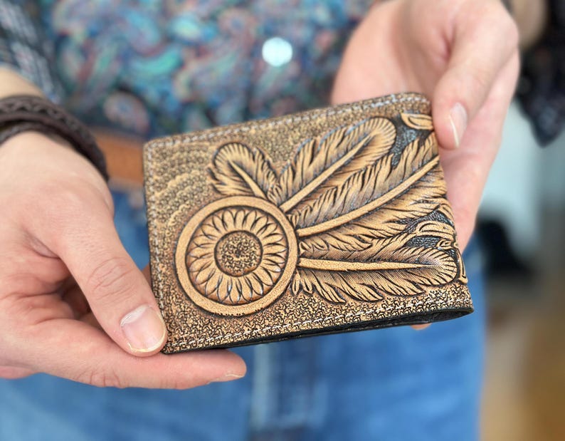 May include: A brown leather wallet with a tooled design of feathers and a dreamcatcher. The wallet has a black stitching around the edges.